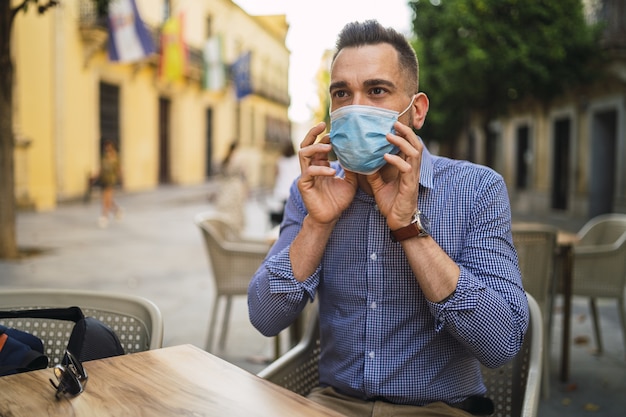 Junger Mann in einem blauen Hemd, das eine medizinische Gesichtsmaske trägt, die in einem Straßencafé - Covid-19-Konzept sitzt