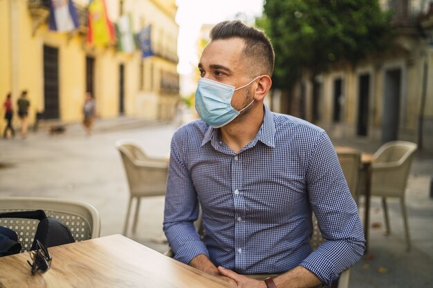 Junger Mann in einem blauen Hemd, das eine medizinische Gesichtsmaske trägt, die in einem Straßencafé sitzt