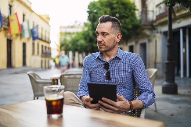 Junger Mann in einem formellen Outfit, das in einem Straßencafé sitzt, das eine Tablette hält und kaltes Getränk trinkt