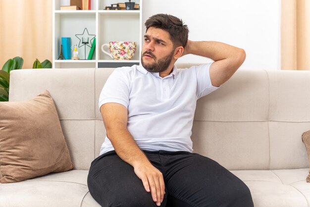 Junger Mann in Freizeitkleidung, der beiseite schaut, verwirrt mit der Hand auf dem Kopf, der auf einer Couch im hellen Wohnzimmer sitzt