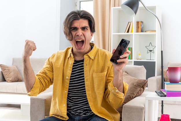 Junger Mann in Freizeitkleidung, der das Smartphone mit der Faust hält verrückt verrückt schreien frustriert auf dem Stuhl im hellen Wohnzimmer sitzen light