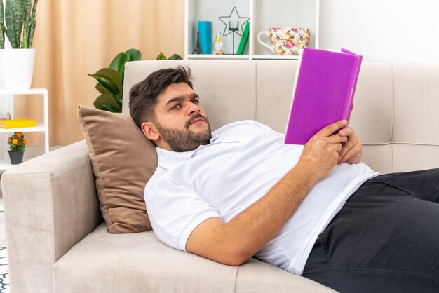 Junger Mann in Freizeitkleidung, der ein Buch mit ernstem Gesicht liest und das Wochenende zu Hause auf einer Couch im hellen Wohnzimmer verbringt