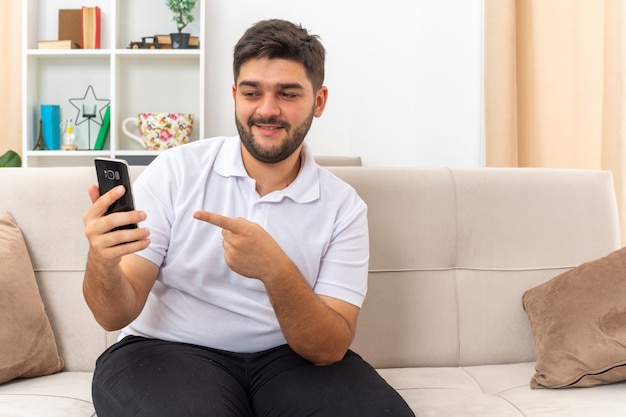 Junger Mann in Freizeitkleidung, der ein Smartphone hält und mit dem Zeigefinger darauf zeigt, lächelt selbstbewusst auf einer Couch im hellen Wohnzimmer sitzen