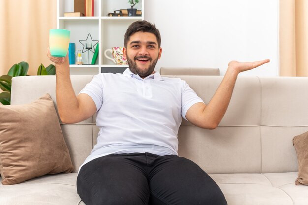 Junger Mann in Freizeitkleidung, der eine Tasse hält, die glücklich und fröhlich aussieht und den Arm zur Seite ausbreitet, der auf einer Couch im hellen Wohnzimmer sitzt