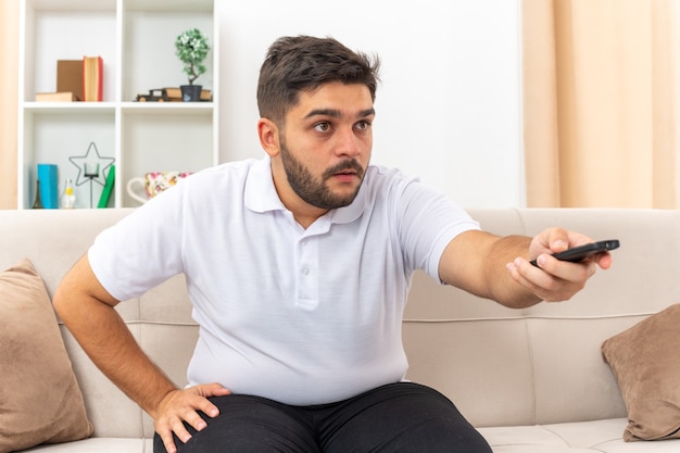 Junger Mann in Freizeitkleidung, der eine TV-Fernbedienung vor dem Fernseher hält und fasziniert schaut, das Wochenende zu Hause auf einer Couch im hellen Wohnzimmer zu verbringen?