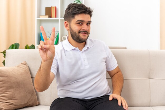 Junger Mann in Freizeitkleidung, der fröhlich lächelt und ein V-Zeichen zeigt, das auf einer Couch im hellen Wohnzimmer sitzt sitting