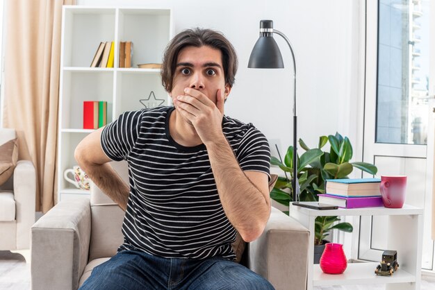 Junger Mann in Freizeitkleidung, der schockiert ist und den Mund mit der Hand bedeckt, die auf dem Stuhl im hellen Wohnzimmer sitzt?