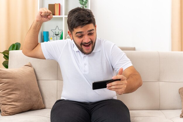 Junger Mann in Freizeitkleidung, der Spiele mit Smartphone-Faustballen spielt, glücklich und aufgeregt auf einer Couch im hellen Wohnzimmer sitzend