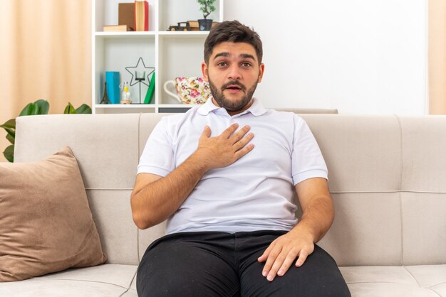 Junger Mann in Freizeitkleidung, der verwirrt aussieht und auf sich selbst zeigt, der auf einer Couch im hellen Wohnzimmer sitzt