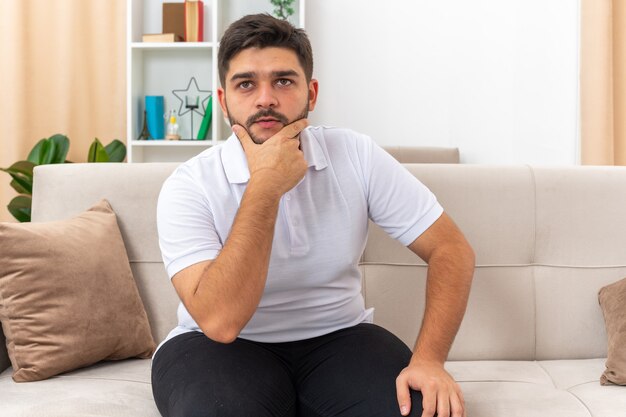 Junger Mann in Freizeitkleidung, der verwirrt mit der Hand am Kinn auf einer Couch im hellen Wohnzimmer sitzt