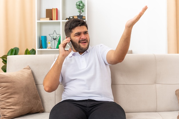 Junger Mann in Freizeitkleidung, der verwirrt und unzufrieden aussieht, während er am Handy spricht und empört den Arm hebt, der auf einer Couch im hellen Wohnzimmer sitzt