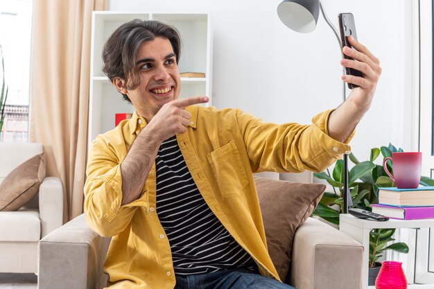 Junger Mann in Freizeitkleidung macht Selfie mit Smartphone glücklich und fröhlich und zeigt mit dem Zeigefinger auf den Bildschirm, der auf dem Stuhl im hellen Wohnzimmer sitzt