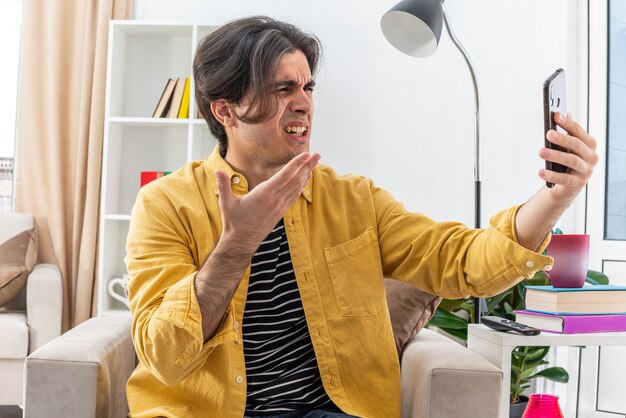 Junger Mann in Freizeitkleidung macht Selfie mit Smartphone und sieht wütend und frustriert auf dem Stuhl im hellen Wohnzimmer aus