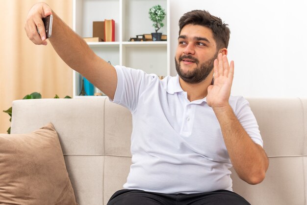 Junger Mann in Freizeitkleidung macht Selfie mit Smartphone winkt mit der Hand und lächelt fröhlich und verbringt das Wochenende zu Hause auf einer Couch im hellen Wohnzimmer sitzend