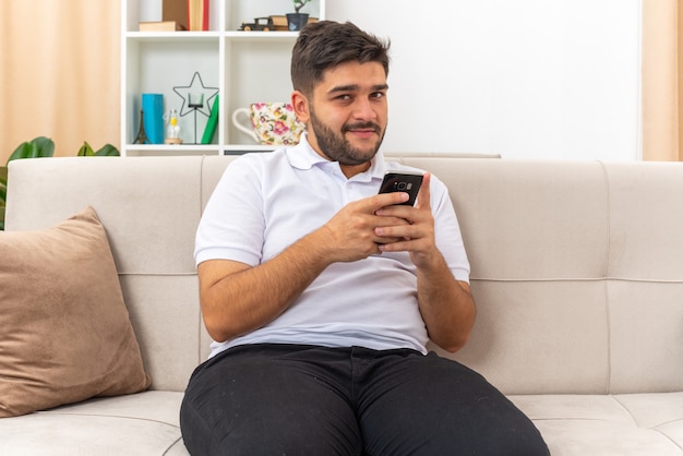 Junger Mann in Freizeitkleidung mit Smartphone, der glücklich und positiv lächelt, zuversichtlich, auf einer Couch im hellen Wohnzimmer sitzend