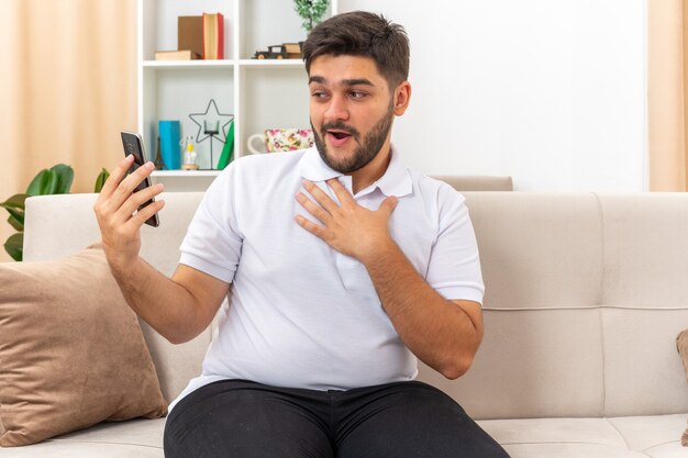 Junger Mann in Freizeitkleidung mit Smartphone mit Videoanruf glücklich und positiv lächelnd auf einer Couch im hellen Wohnzimmer sitzend