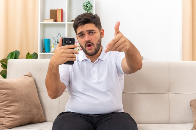 Junger Mann in Freizeitkleidung mit Smartphone sieht glücklich und selbstbewusst auf einer Couch im hellen Wohnzimmer aus