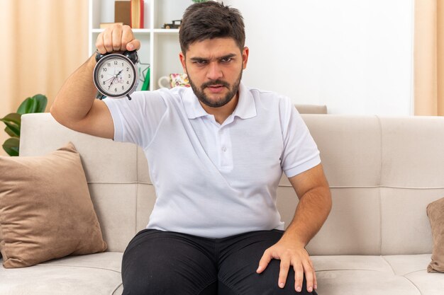 Junger Mann in Freizeitkleidung mit Wecker, der mit ernstem Stirnrunzeln auf einer Couch im hellen Wohnzimmer sitzt