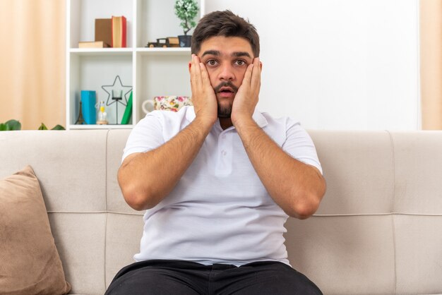Junger Mann in Freizeitkleidung sieht erstaunt und überrascht aus, das Wochenende zu Hause auf einer Couch im hellen Wohnzimmer zu verbringen living