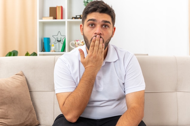 Junger Mann in Freizeitkleidung sieht erstaunt und überrascht aus, den Mund mit der Hand zu bedecken, die auf einer Couch im hellen Wohnzimmer sitzt