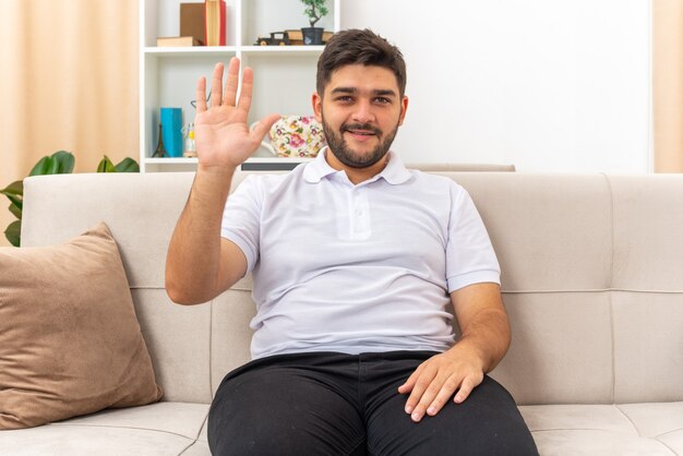 Junger Mann in Freizeitkleidung sieht glücklich und selbstbewusst aus und winkt mit der Hand, die auf einer Couch im hellen Wohnzimmer sitzt