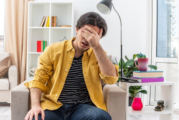 Junger Mann in Freizeitkleidung sieht müde und gelangweilt aus und bedeckt die Augen mit der Hand, die auf dem Stuhl im hellen Wohnzimmer sitzt