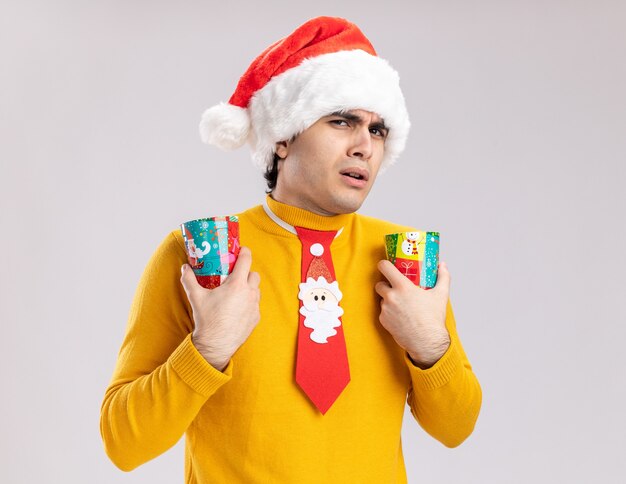 Junger Mann in gelbem Rollkragenpullover und Weihnachtsmütze mit lustiger Krawatte, die bunte Pappbecher hält, die Kamera verwirrt und unzufrieden stehen über weißem Hintergrund betrachten