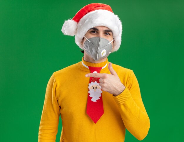 Junger Mann in gelbem Rollkragenpullover und Weihnachtsmütze mit lustiger Krawatte, die Gesichtsschutzmaske trägt, die Kamera mit glücklichem Gesicht betrachtet, das mit Zeigefinger zur Seite steht, die über grünem Hintergrund steht