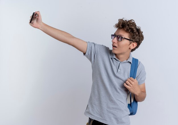 Junger Mann in grauem Poloshirt, das Smartphone hält, das Selfie lächelnd über weißer Wand steht