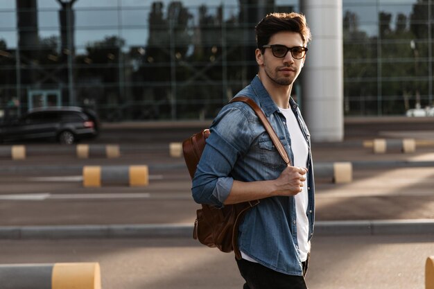 Junger Mann in Jeansjacke mit Sonnenbrille und weißem T-Shirt schaut in die Kamera Schöner brünetter Kerl hält Rucksack und posiert draußen