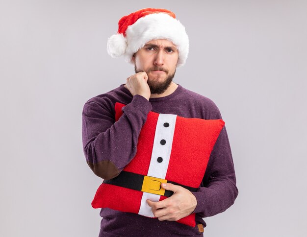 Junger Mann in lila Pullover und Weihnachtsmütze mit lustigem Kissen und Blick in die Kamera unzufrieden auf weißem Hintergrund stehend