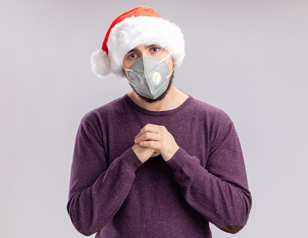 Junger Mann in lila Pullover und Weihnachtsmütze tragen Gesichtsschutzmaske Händchenhalten zusammen mit Blick auf Kamera müde und gelangweilt stehend über weißem Hintergrund