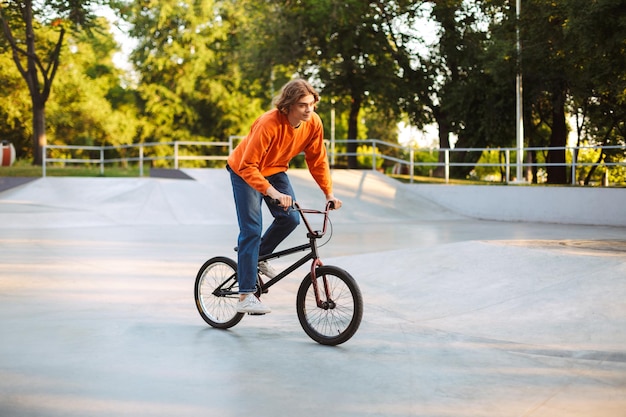 Junger Mann in orangefarbenem Pullover und Jeans fährt verträumt Fahrrad im modernen Skatepark