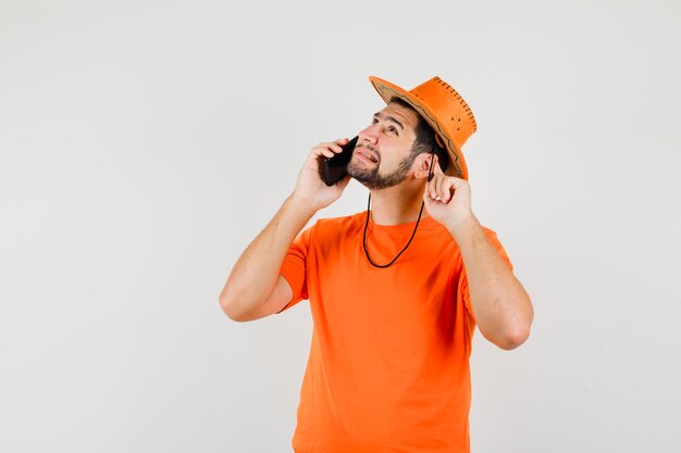 Junger Mann in orangefarbenem T-Shirt, Hut, der mit dem Handy spricht und Probleme mit dem Hören hat, Vorderansicht.