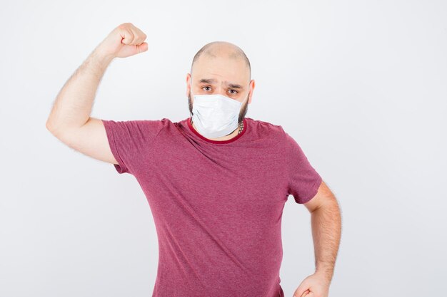 Junger Mann in rosa T-Shirt, Maske, die seine Armmuskeln zeigt und flexibel aussieht, Vorderansicht.