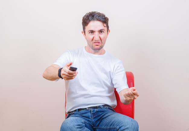 Junger Mann in T-Shirt, Jeans mit Fernbedienung, während er auf einem Stuhl sitzt und wütend aussieht