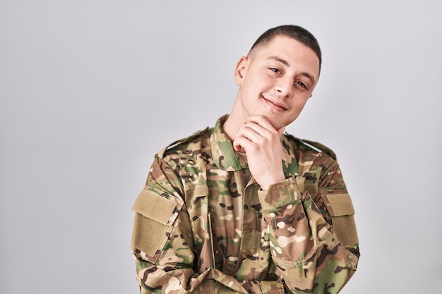 Junger Mann in Tarnuniform der Armee blickt selbstbewusst in die Kamera und lächelt mit verschränkten Armen und erhobener Hand am Kinn. positiv denken.