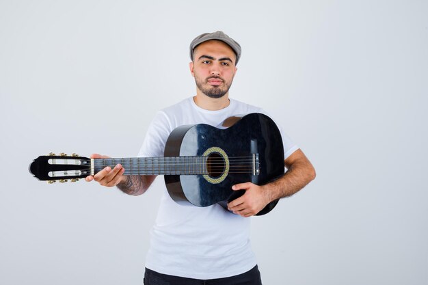 Junger Mann in weißem T-Shirt, schwarzer Hose, grauer Mütze, der Gitarre spielt und ernst aussieht