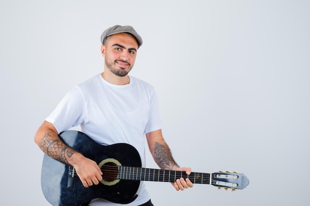 Junger Mann in weißem T-Shirt, schwarzer Hose, grauer Mütze, der Gitarre spielt und glücklich aussieht