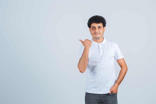 Junger Mann in weißem T-Shirt und Jeans, der die Hand in die Tasche steckt, während er weg zeigt und ernst aussieht, Vorderansicht.