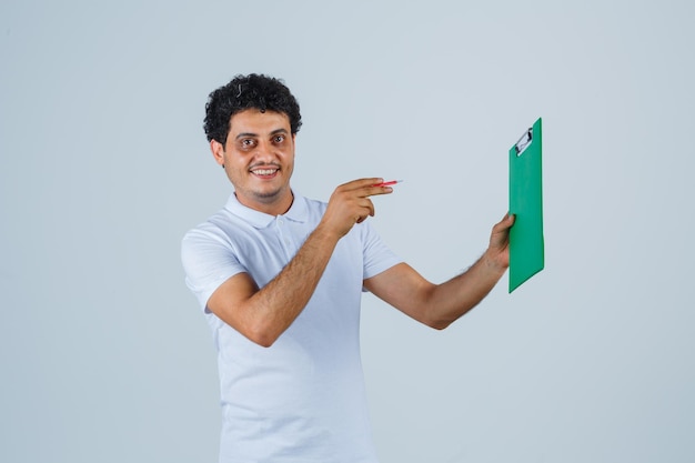 Junger Mann in weißem T-Shirt und Jeans, der ein Notizbuch hält und mit einem Stift darauf zeigt, in die Kamera schaut und glücklich aussieht, Vorderansicht.