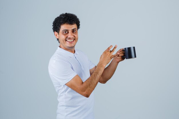 Junger Mann in weißem T-Shirt und Jeans, der eine Tasse Tee hält, während er darauf zeigt und glücklich aussieht, Vorderansicht.