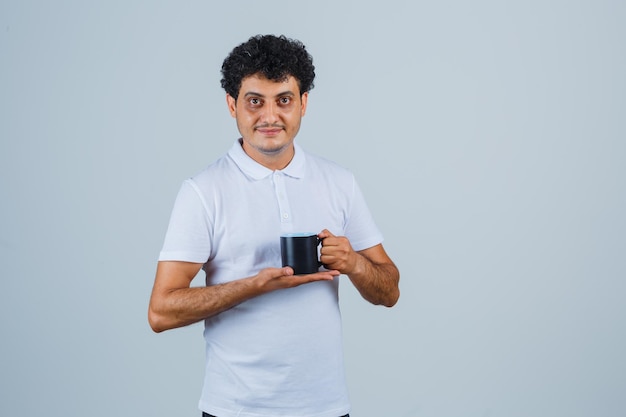 Junger Mann in weißem T-Shirt und Jeans, der mit beiden Händen eine Tasse Tee hält und glücklich aussieht, Vorderansicht.