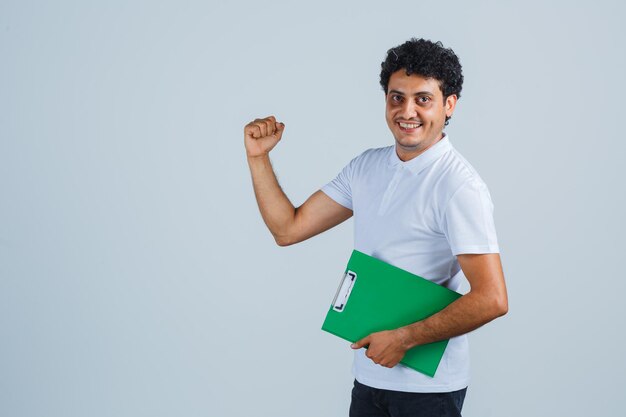 Junger Mann in weißem T-Shirt und Jeans, der Notizbuch hält und glücklich geht und schaut, Vorderansicht.