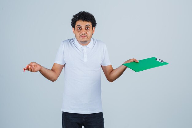 Junger Mann in weißem T-Shirt und Jeans, der Notizbuch und Stift hält, die Schultern zuckt und verwirrt aussieht, Vorderansicht.