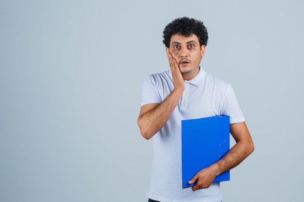 Junger Mann in weißem T-Shirt und Jeans mit Aktenordner, Hand in der Nähe des Mundes und überrascht, Vorderansicht.