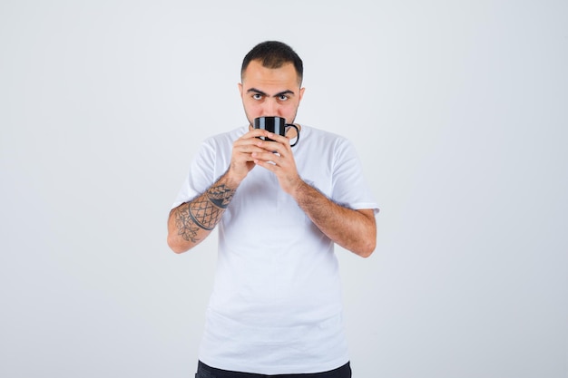 Junger Mann in weißem T-Shirt und schwarzer Hose, der eine Tasse Tee hält und ihn trinkt und ernst aussieht