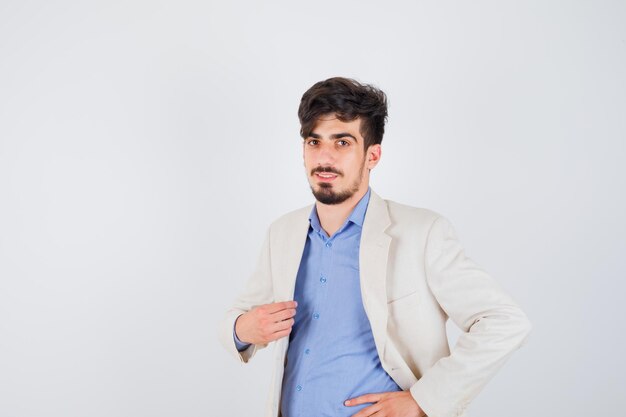Junger Mann legt eine Hand auf die Jacke, eine andere Hand auf die Taille in blauem Hemd und weißer Anzugjacke und sieht selbstbewusst aus