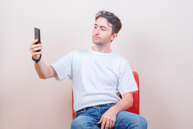 Junger Mann macht Selfie auf dem Handy, während er auf einem Stuhl in T-Shirt, Jeans sitzt
