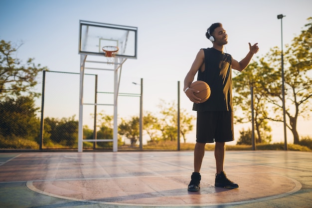 Junger mann macht sport, spielt basketball bei sonnenaufgang, hört musik über kopfhörer